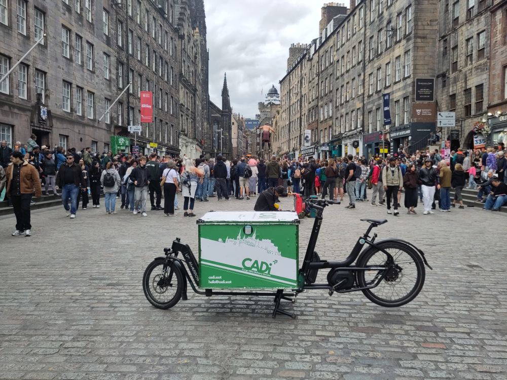 Urban Cargo XL bike with Cadi Couriers coop branding, in front a crowd of people on the High Street near the cast in Edinburgh.
