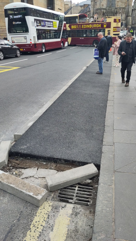 Extended pavement for pedestrians next to a busy road filled with buses. The extended pavement abruptly ends near the bottom of the image, having clearly been cut away to uncover a drain in the street, with bits of broken concrete blocks around it.