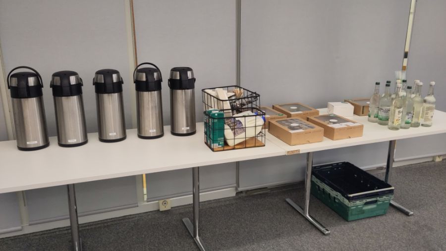 A long table with 5 hot drink flasks, two crates containing milk, sugar, cups and stirrers on the left. To the right of the crates are four meal platters, 10 bottles of still and sparkling water, and an additional meal platter partially obscured behind the water bottles.