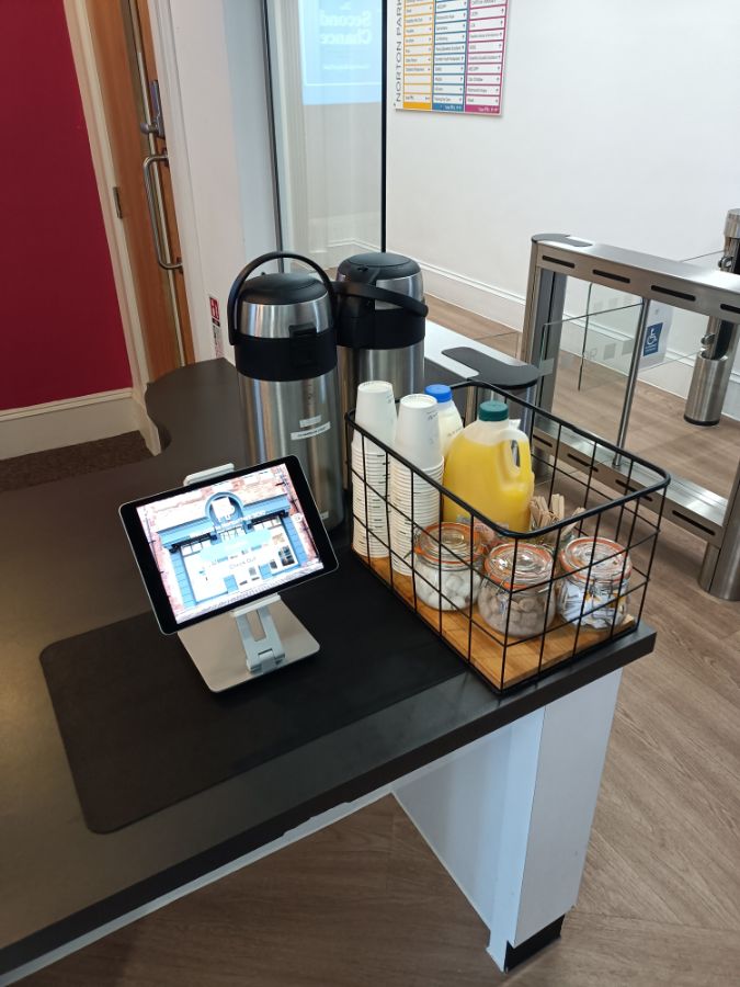 A reception area with admission gates to one side. Two flasks of hot drinks sit on the reception counter. In front of the flasks is a wire crate with disposable cups, sugar, stirrers, orange juice and milk.